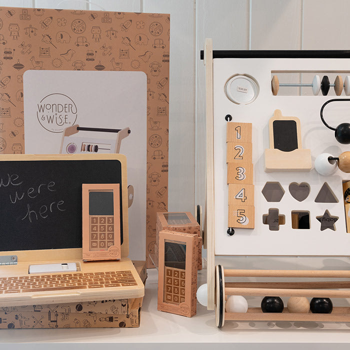 A display of wooden and cardboard educational toys, including a laptop, cellphones, an activity board with numbers, shapes, and abacus, set against a box labeled
