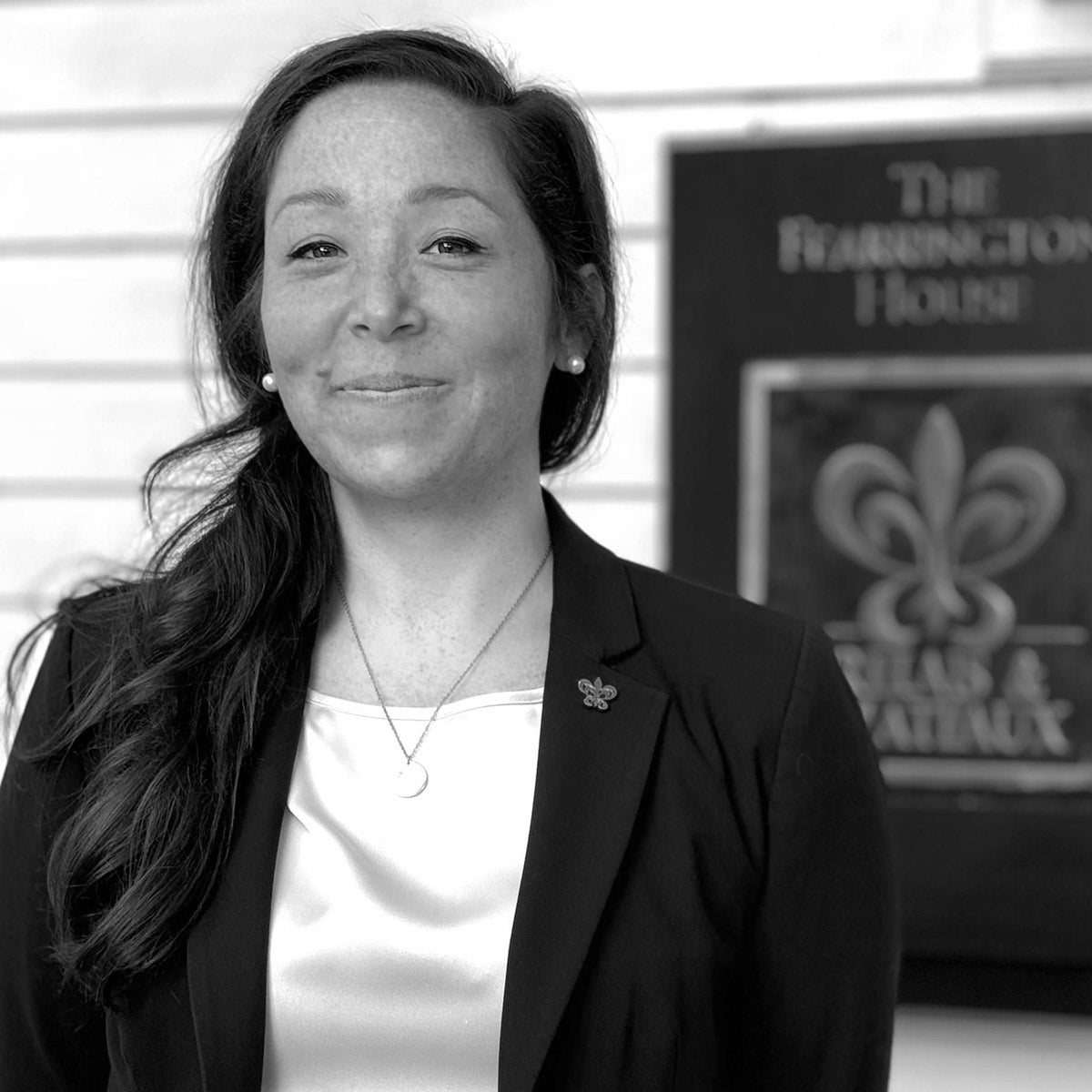 A woman with long dark hair, wearing a blazer and light blouse, stands smiling in front of a sign that reads “The Harrington House.” The photo is in black and white.