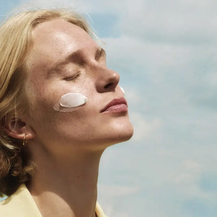 A person with closed eyes faces the sun, with a smear of white cream on their cheek. They have light skin, blond hair, and are outdoors under a partly cloudy sky.