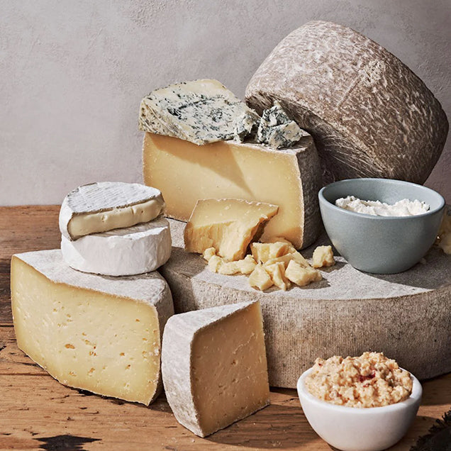 A variety of cheeses, including wheels, wedges, and rounds, are arranged on a wooden surface with bowls of crumbled cheese and spread, creating a rustic cheese platter display.