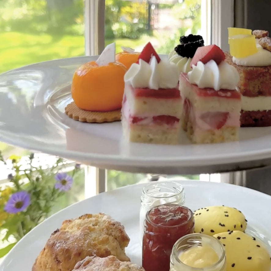 Two plates with assorted pastries near a sunny window: one plate has fruit-topped cakes, a tart, and a round orange dessert; the other has scones, jams in small jars, and yellow macarons with poppy seeds. Purple flowers are outside.