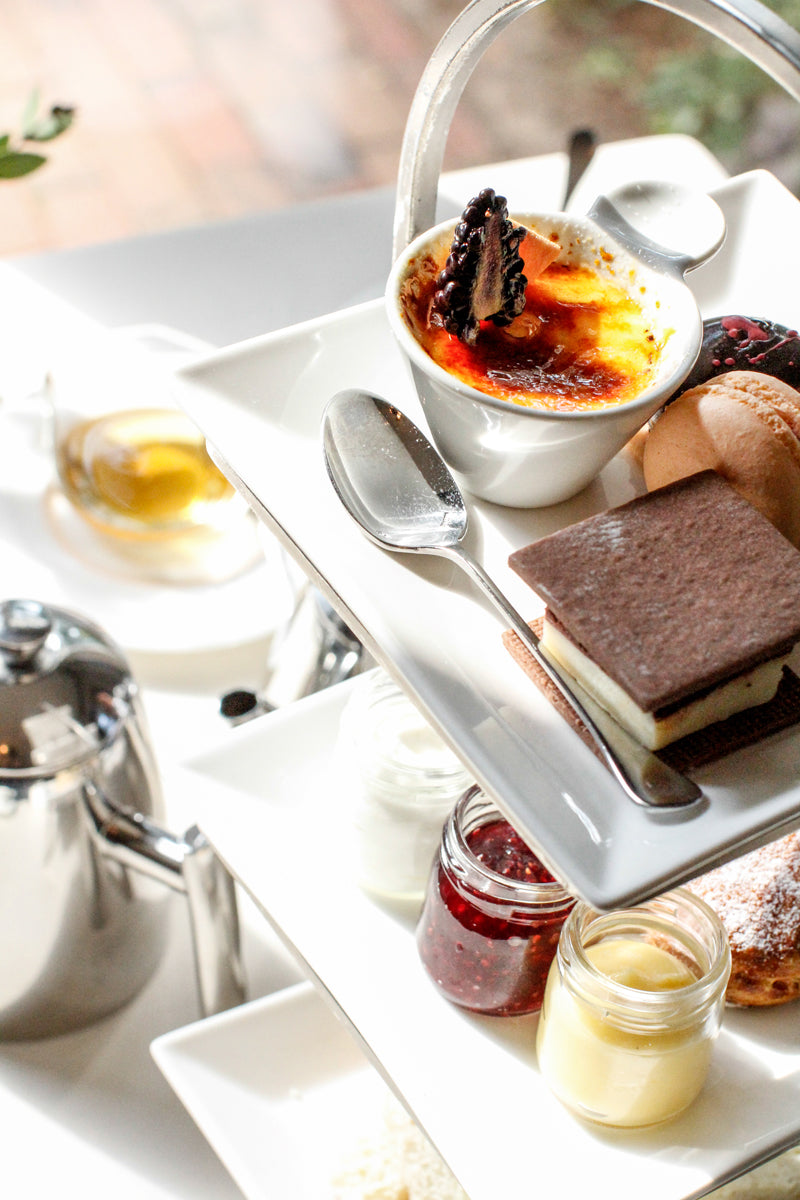 A tiered tray with assorted desserts, including crème brûlée, layered cake, jams, and cream, sits on a white table next to a teapot and a cup of tea, all brightly lit by natural light.