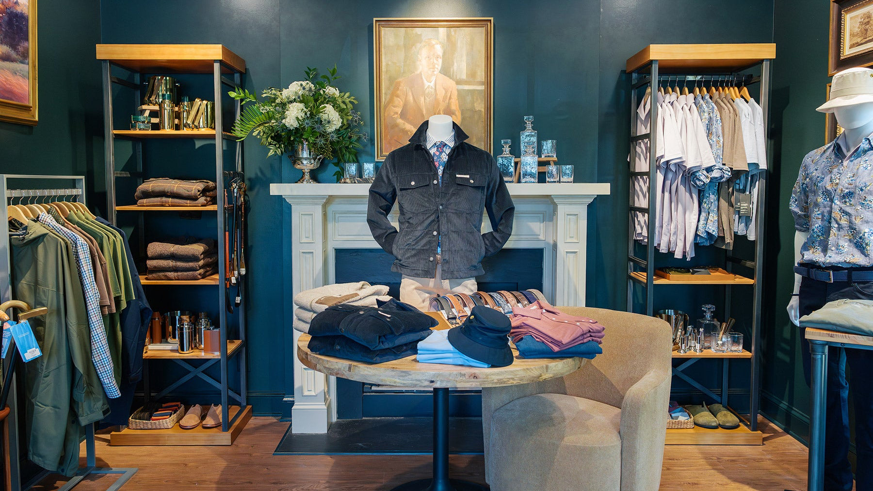 A stylish clothing store interior with shirts, jackets, and pants neatly displayed on racks and a round table. A cozy armchair sits nearby, and a portrait hangs above a white fireplace with decorative items.