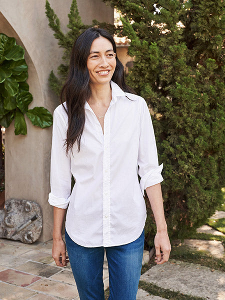 A woman with long dark hair wearing a white button-up shirt and blue jeans stands outdoors, smiling, with greenery and a stone path in the background.
