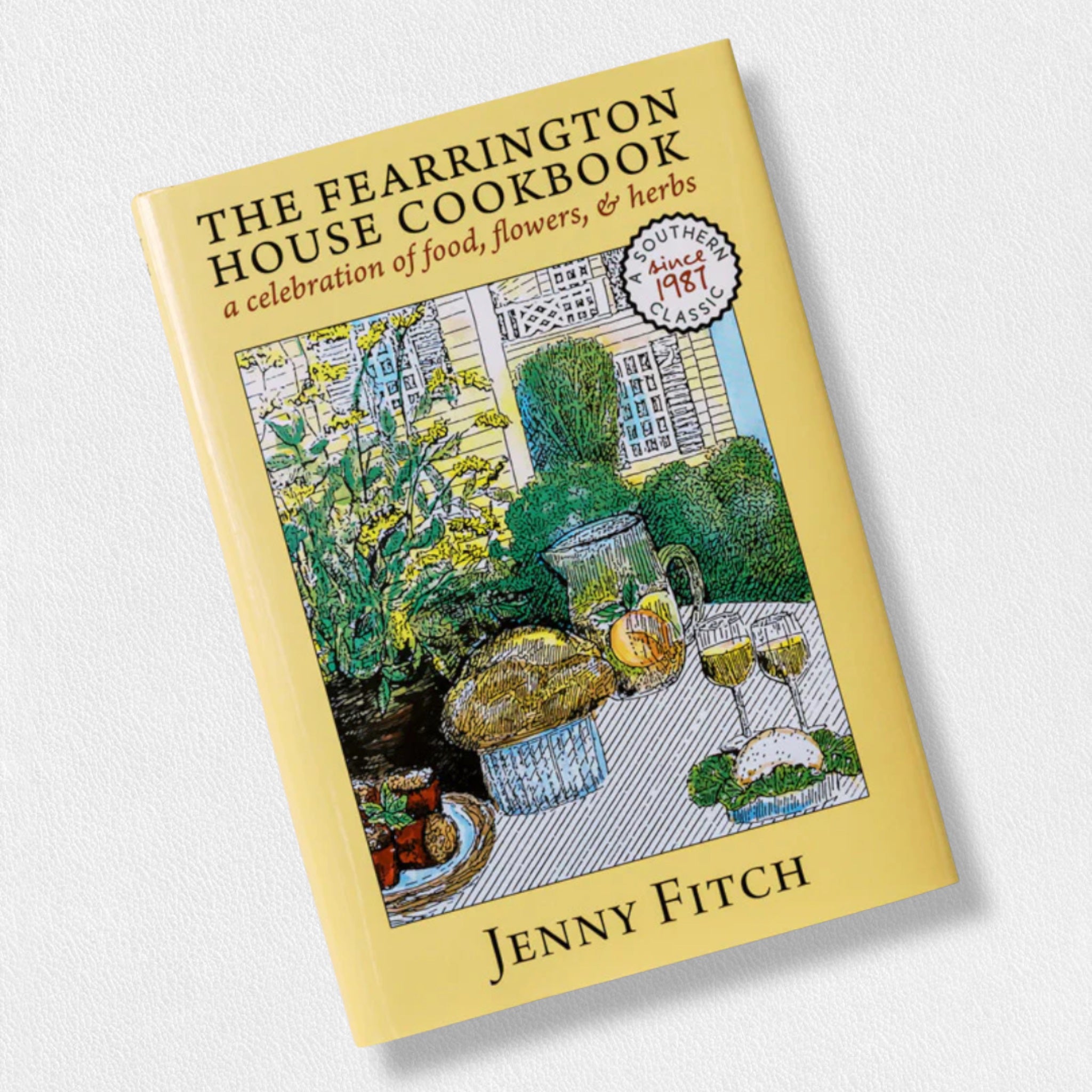 The FEARRINGTON SIGNATURE COLLECTION – THE FEARRINGTON HOUSE COOKBOOK BY JENNY FITCH from BELTED GOAT features an illustrated cover with an outdoor table set with food, wine, and flowers against a yellow border—celebrating Southern cuisine and hospitality.