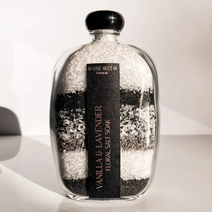A glass bottle containing layers of coarse, white, and black bath salts is displayed against a light background. The bottle features a black label reading