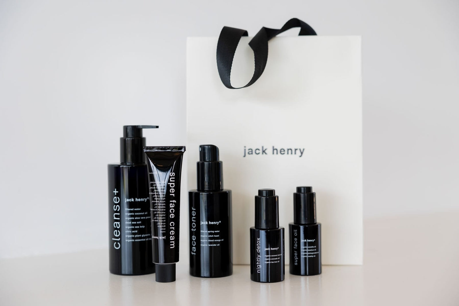 Five black Jack Henry skincare bottles in front of a white Jack Henry shopping bag.