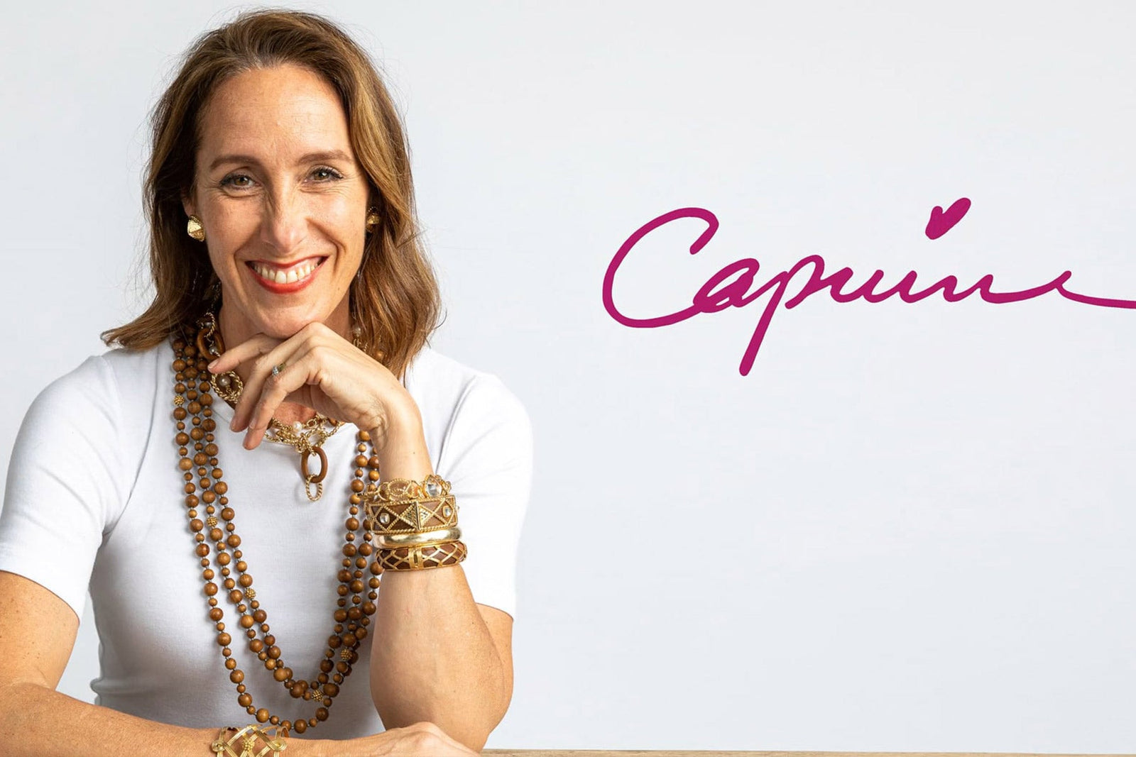 Portrait of a smiling woman wearing layered gold and beaded necklaces and stacked bracelets, with the Capucine logo in pink script beside her.