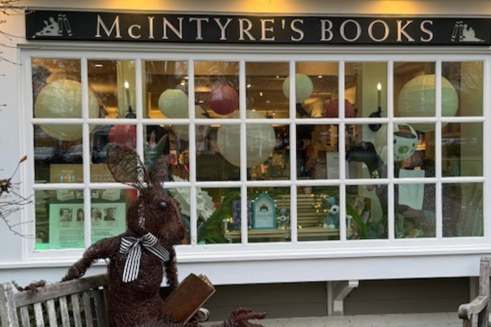 Storefront of McIntyre’s Books with large paned windows glowing warmly inside. In front, a wire bunny sculpture with a striped bow sits on a bench holding an open book, creating a whimsical, storybook-style scene outside the shop.