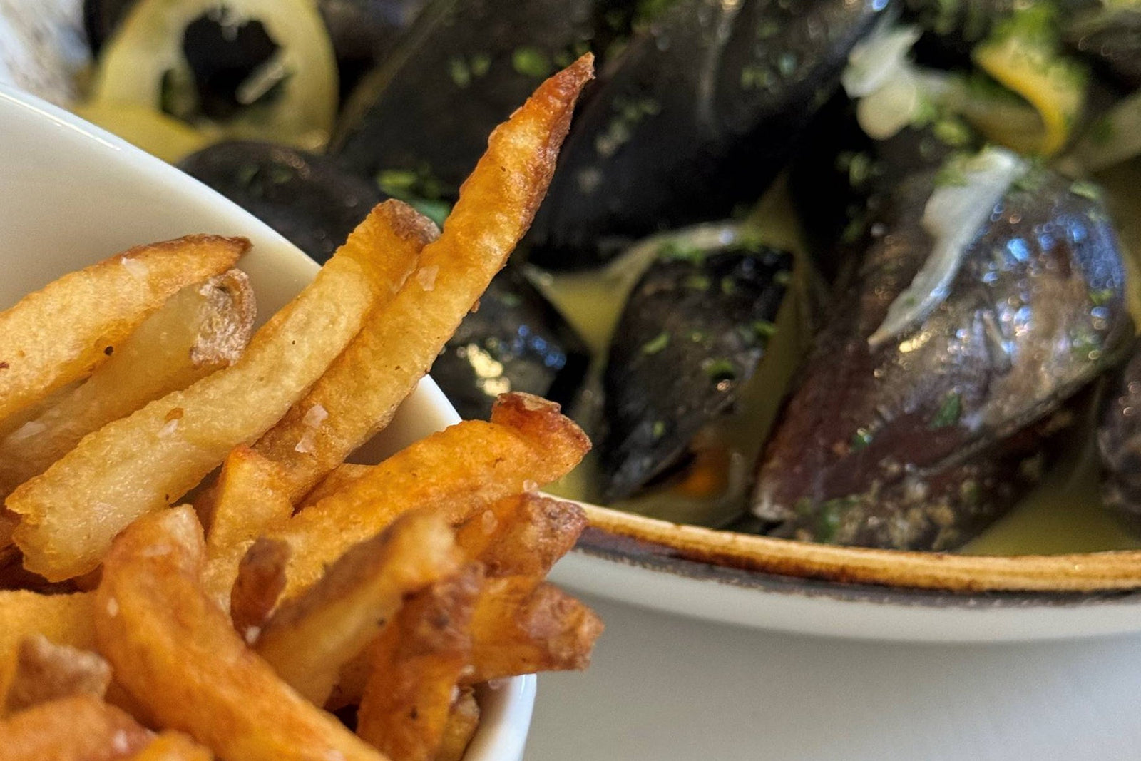 A close-up of crispy French fries in a white cup beside a bowl of mussels in a herb-butter broth.