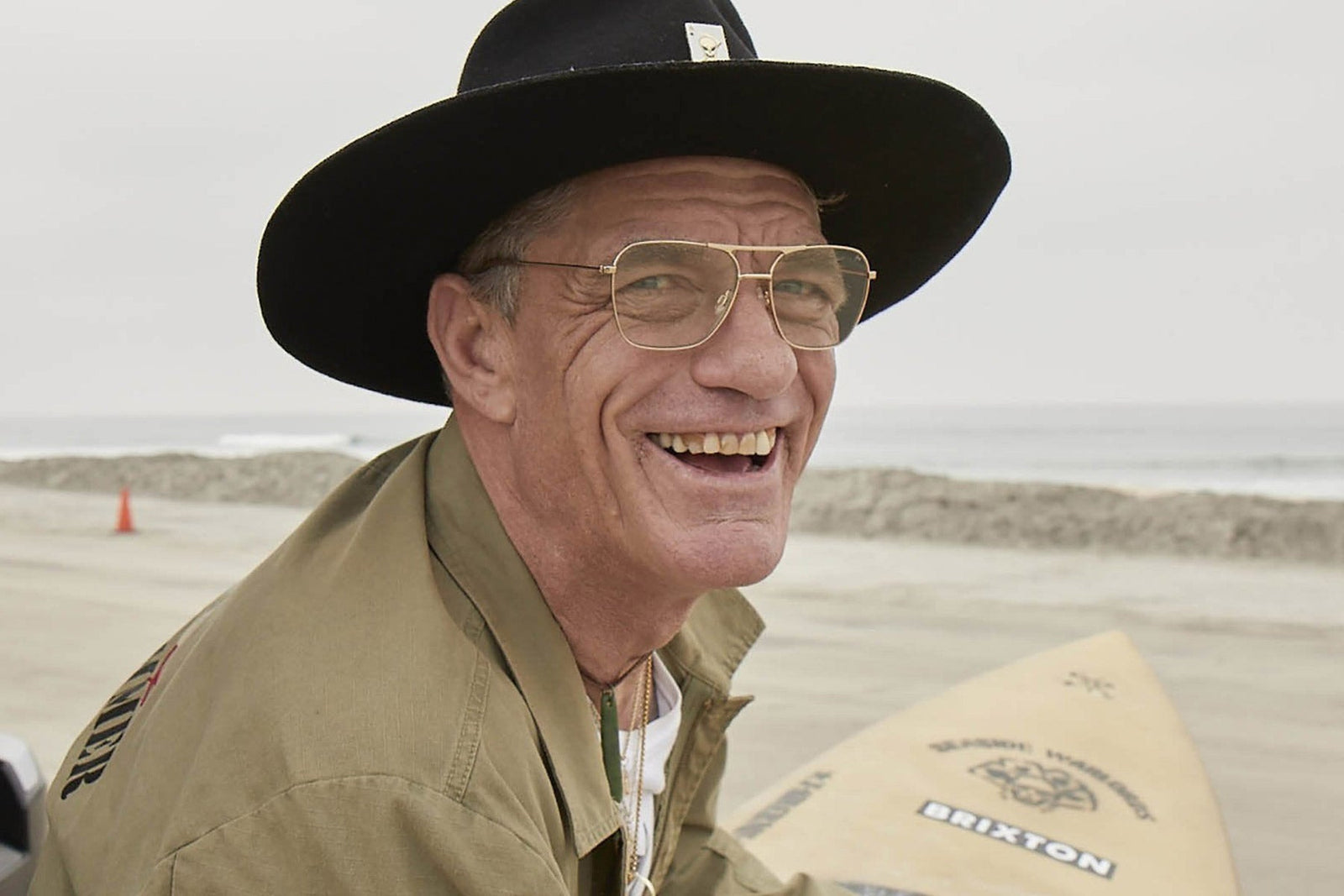 Smiling man wearing a black wide-brim hat and glasses sits on a beach beside a surfboard, with the ocean in the background.