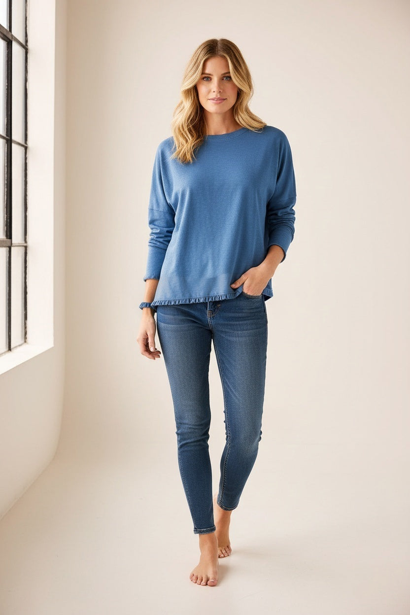 A woman stands barefoot indoors near a large window, looking at the camera with a relaxed expression. She wears the MERSEA - Catalina Ruffle Tee in Steel Blue by MERSEA, featuring delicate feminine ruffle details, paired with blue skinny jeans. Her blonde hair falls loosely around her shoulders.