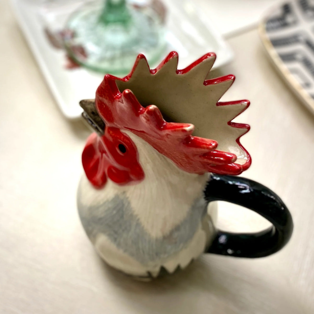 The QUAIL CERAMICS - DORKING ROOSTER JUG by QUAIL DESIGNS, a hand-painted stoneware jug with a red comb and black handle, sits on the table beside a green glass and patterned plates—a charming British tea accessory.