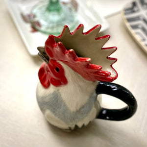 The QUAIL CERAMICS - DORKING ROOSTER JUG by QUAIL DESIGNS, a hand-painted stoneware jug with a red comb and black handle, sits on the table beside a green glass and patterned plates—a charming British tea accessory.