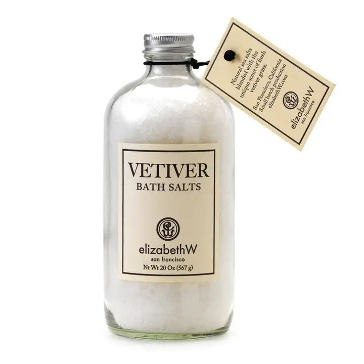 A clear glass bottle labeled "VETIVER BATH SALTS" from E WIGHTMAN &amp; CO, San Francisco, contains natural white bath salts. The bottle features a silver metal cap and a rectangular tag attached with black string, offering product information and highlighting the amazing aroma for a soothing bath experience.