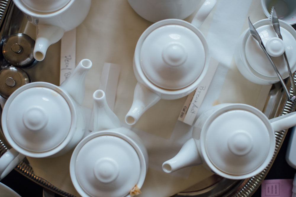 Overhead view of several white teapots with lids on a tray, arranged closely together. Some have handwritten labels underneath, and there are also salt and pepper shakers visible in the corner.