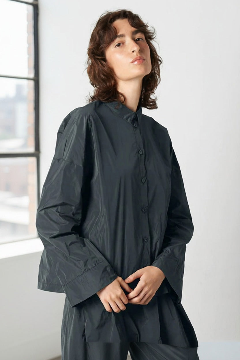 A person with wavy brown hair wears the KATHARINA HOVMAN - KIMONO BLOUSE by KATHARINA HOVMAN in dark gray, standing against a plain light background and looking slightly upward with a neutral expression.