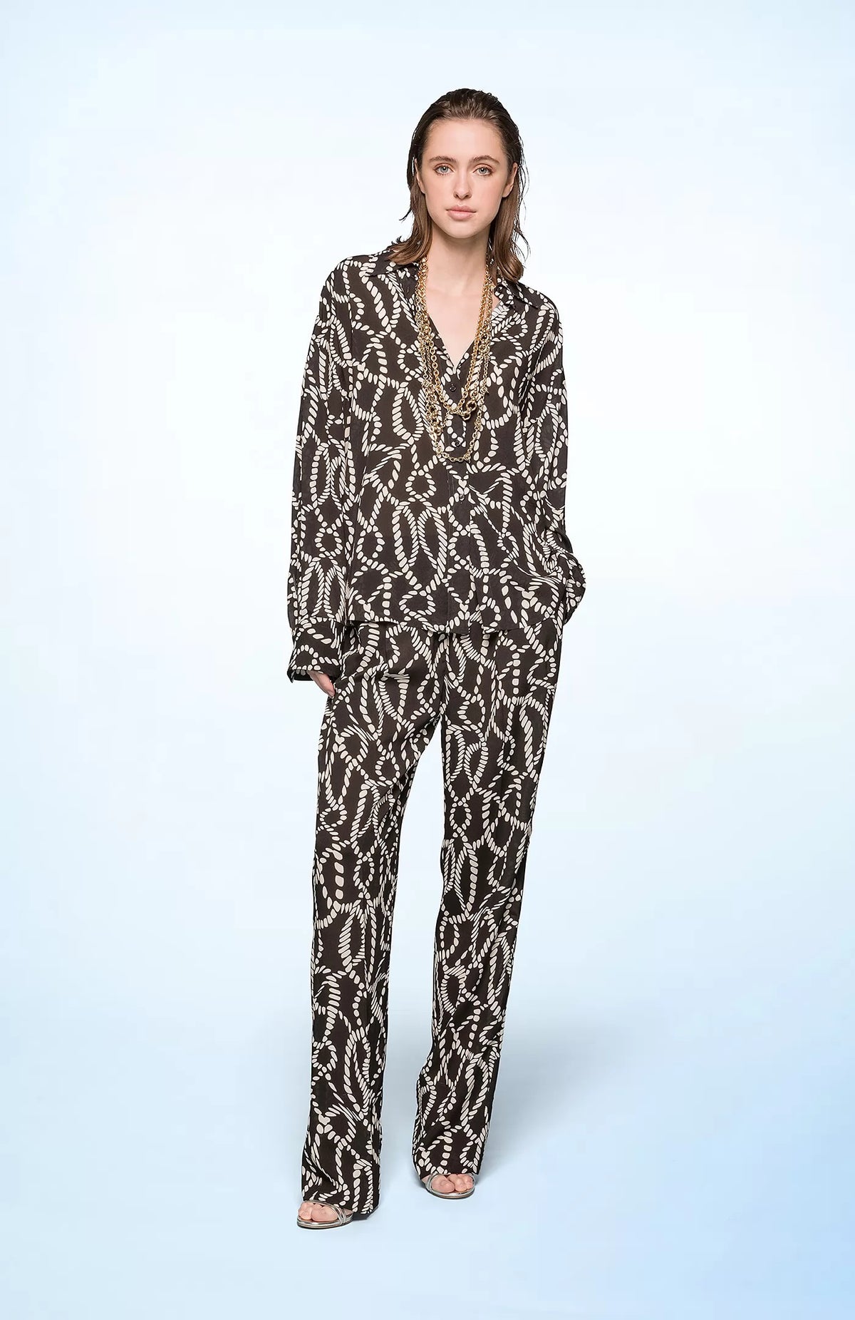 A person with light skin and shoulder-length brown hair stands against a light blue background. They are wearing the SEVENTY VENEZIA - PRINTED SHIRT WITH OPEN BACK by SEVENTY, a loose-fitting black and white patterned outfit made of lightweight fabric. The person gazes over their shoulder towards the camera, showcasing the open back detail of the shirt.