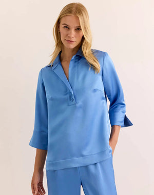 A woman with blonde hair wears the HINSON WU Aileen 3/4 Sleeve Liquid Satin Shirt in Sky Blue—a loose-fitting blouse featuring an elegant drape and luminous sheen. Paired with matching pants, the light blue satin fabric from HINSON WU catches the light beautifully as she stands against a simple, light background.