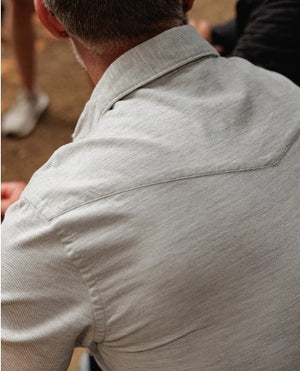 Close-up of a person wearing the CRIQUET Long Sleeve Corduroy Pearl Snap Shirt in Heather Gray with the top buttons open, revealing some chest hair. The shirt features a collar and a breast pocket on the right side.