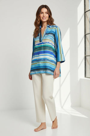 A woman wearing the HINSON WU VICKY 3/4 Sleeve Tencel Textured Stripe Print Top in blue, green, and white stripes with off-white pants stands smiling against a plain background, her light brown hair styled with hoop earrings.