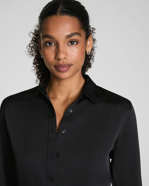 A woman with curly hair pulled back wears the SPANX Silky Smooth Button Down Shirt in black, paired with black pants. She stands facing away, looking over her shoulder against a plain light gray background.