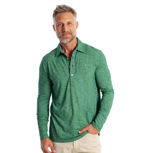 A man with short gray hair and a beard, wearing the CRIQUET - BRRR° Long Sleeve Range Polo in Reed and light pants, stands in a doorway holding a strap with golf clubs and accessories over his shoulder.