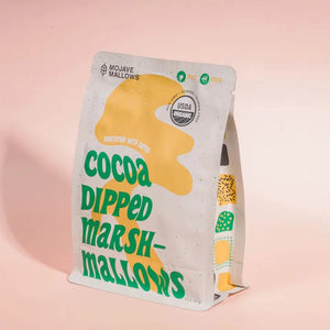A white upright bag of MOJAVE MALLOWS - COCOA DIPPED MARSHMALLOWS by MOJAVE MALLOWS features green and yellow text, a bright yellow graphic, and a USDA organic label on the front, highlighting this delicious handmade treat. Each marshmallow is infused with organic vanilla and coated in rich dark cocoa powder for an irresistible snack.