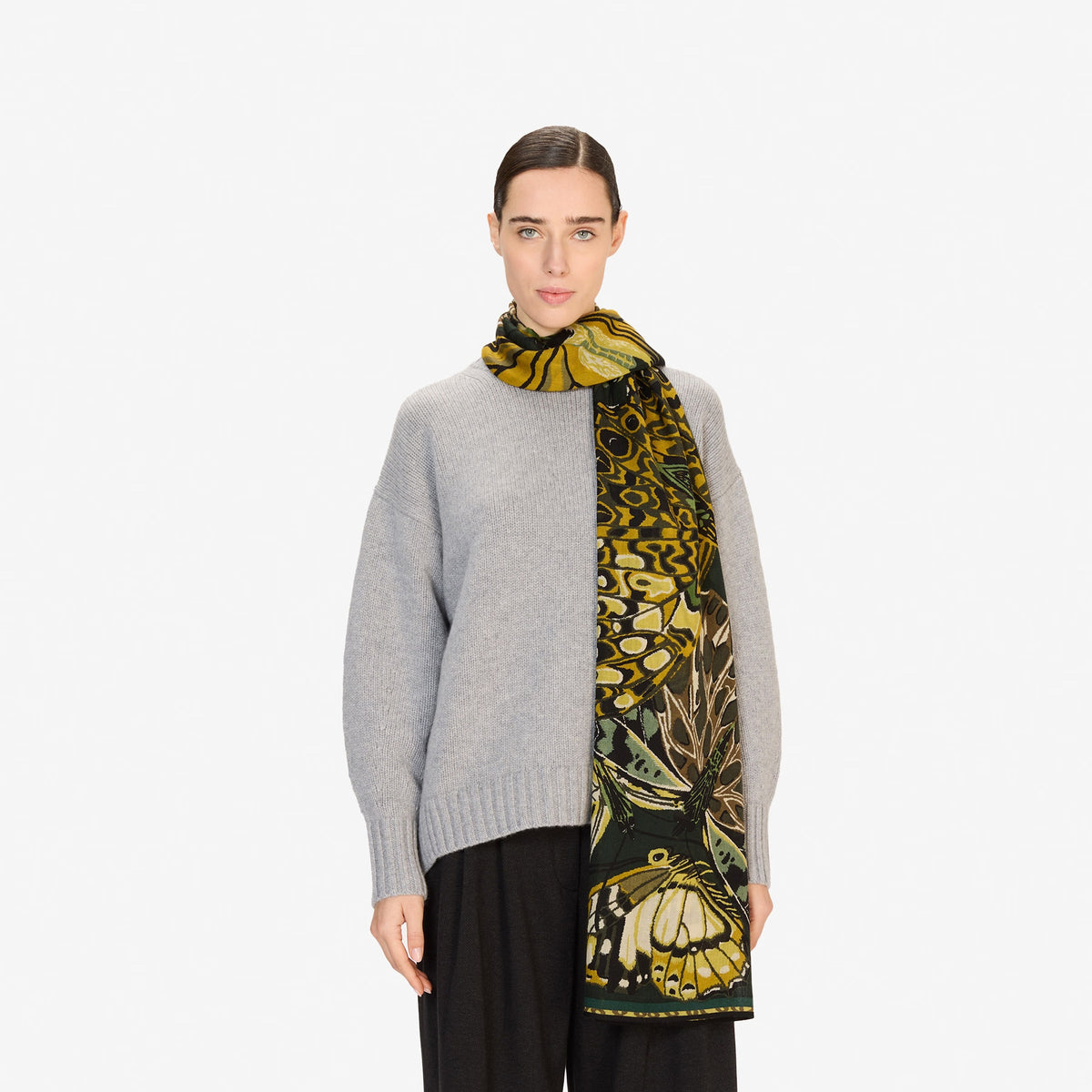 A young man wears the INOUI EDITIONS - PAPILLON (BUTTERFLIES) wool scarf in emerald by INOUI, featuring butterfly and leopard patterns, hand-printed in India, paired with a black shirt, standing against a white background.