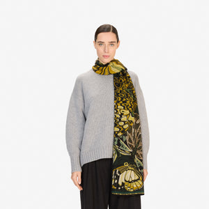 A young man wears the INOUI EDITIONS - PAPILLON (BUTTERFLIES) wool scarf in emerald by INOUI, featuring butterfly and leopard patterns, hand-printed in India, paired with a black shirt, standing against a white background.