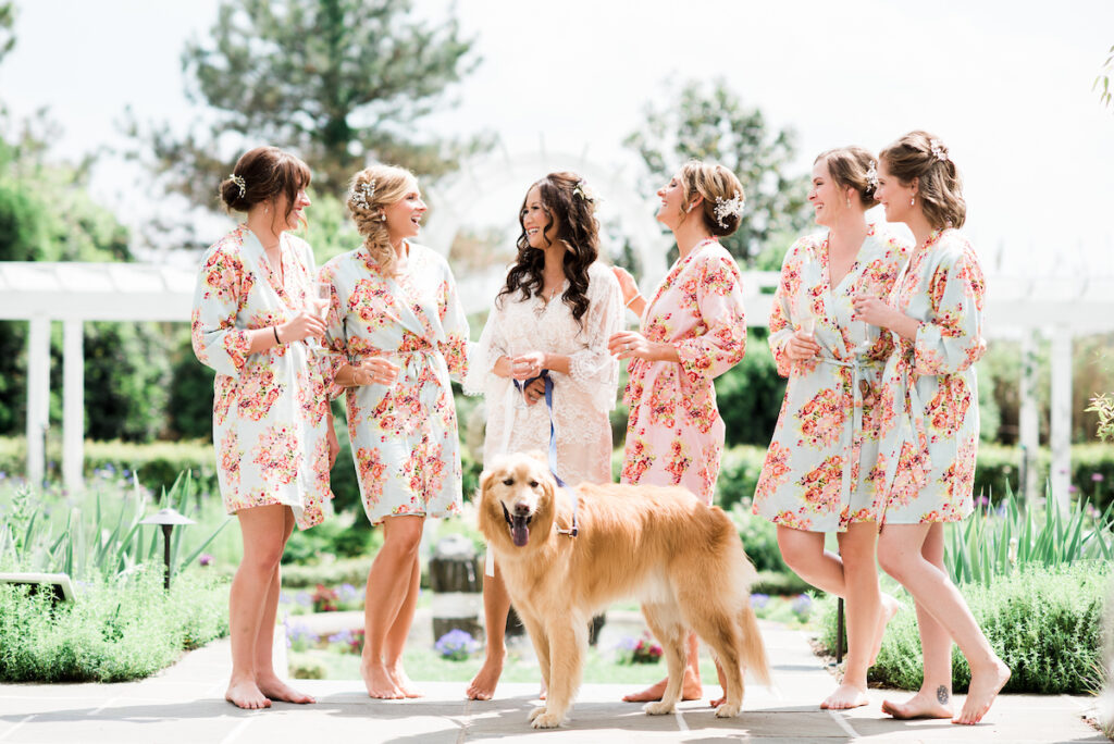 Six women in floral robes, smiling and talking outdoors in a garden. One woman, possibly a bride, stands in the center wearing white. A golden retriever stands in front of them, looking at the camera.