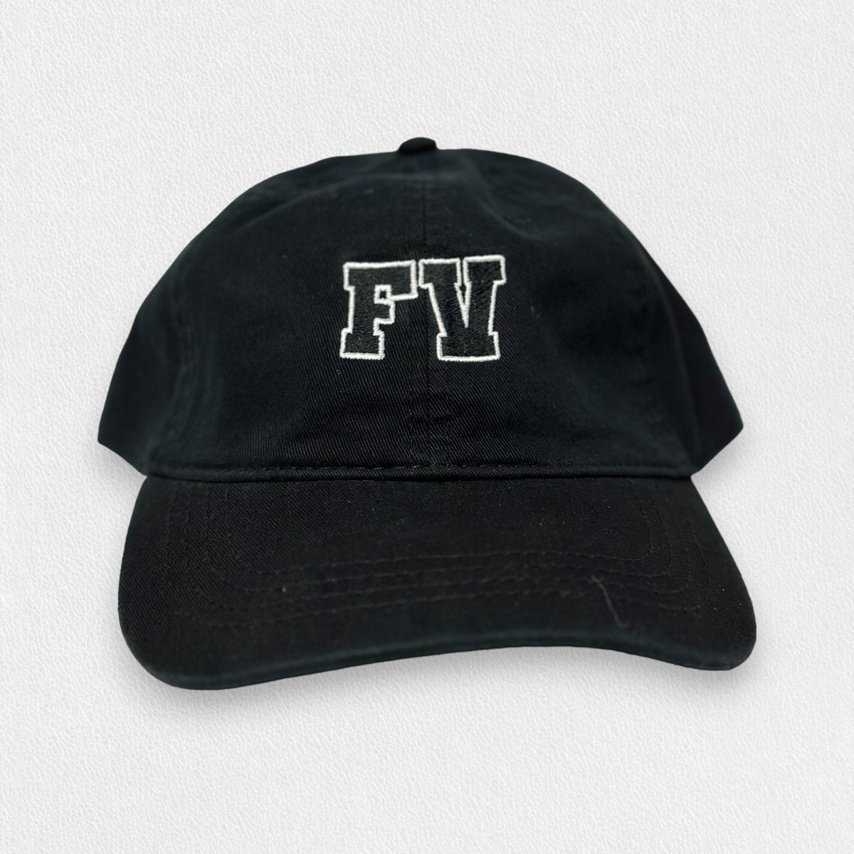 A black &quot;FEARRINGTON LIFESTYLE COLLECTION - ADULT TWILL FV HAT&quot; by STRAN, featuring the letters &quot;FV&quot; embroidered in white on the front. The cap is made from premium cotton twill and includes a slightly curved brim, shown against a plain white background.