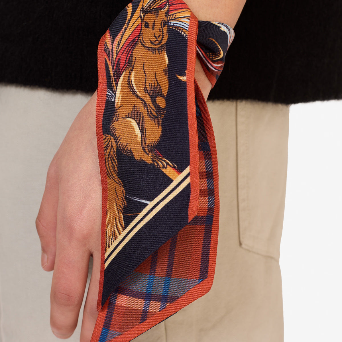 The INOUI EDITIONS - ROCOCO SQUIRREL SILK RIBBON by INOUI is a double-sided silk scarf from India: one side orange and blue plaid, the other features a squirrel and plants on navy with an orange border—an elegant accessory for any wardrobe.