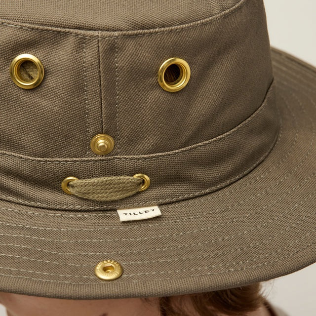 A young person with light skin and wavy brown hair wears the TILLEY - CLASSIC T3 HAT IN OLIVE and a beige crew neck shirt, looking seriously at the camera against a plain light background.