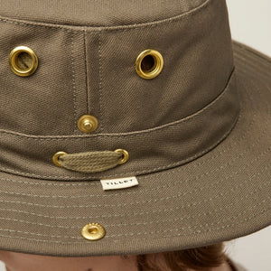 A young person with light skin and wavy brown hair wears the TILLEY - CLASSIC T3 HAT IN OLIVE and a beige crew neck shirt, looking seriously at the camera against a plain light background.