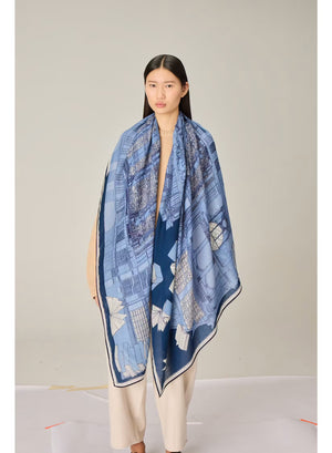 A woman stands against a plain background, wearing a beige sweater, cream wide-leg pants, black shoes, and the INOUI EDITIONS - VIVIENNE WOOL SCARF IN BLUE by INOUI—a long blue scarf with a white pattern made from 100% wool in India and measuring 100 x 190 cm.