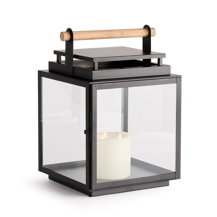 The NAPA HOME AND GARDEN ADWIN OUTDOOR LANTERN features a unique black metal and glass design with a teak handle. Inside, an unlit large white candle enhances its simple and modern appeal.