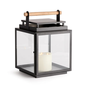 The NAPA HOME AND GARDEN ADWIN OUTDOOR LANTERN features a unique black metal and glass design with a teak handle. Inside, an unlit large white candle enhances its simple and modern appeal.
