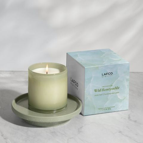 A light green soy wax candle in a hand-blown glass container is placed next to its packaging box. The box, with a watercolor design featuring shades of green and white, displays the text &quot;LAFCO New York, Greenhouse, Wild Honeysuckle.