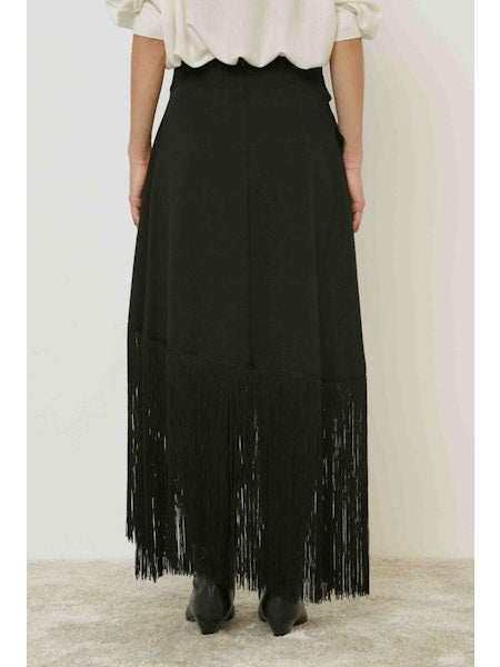 A person stands against a plain background wearing the MEIMEIJ - FRINGE WRAP SKIRT with an asymmetric hem, paired with a light top and black boots.