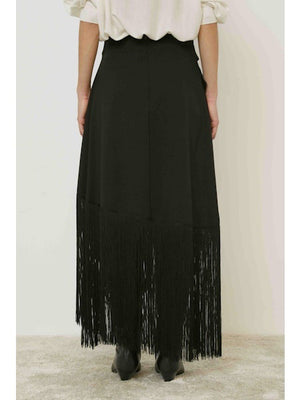 A person stands against a plain background wearing the MEIMEIJ - FRINGE WRAP SKIRT with an asymmetric hem, paired with a light top and black boots.