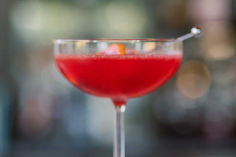 A coupe glass filled with a bright red cocktail, garnished with an orange peel and a cocktail pick, sits on a light-colored surface with a blurred background.