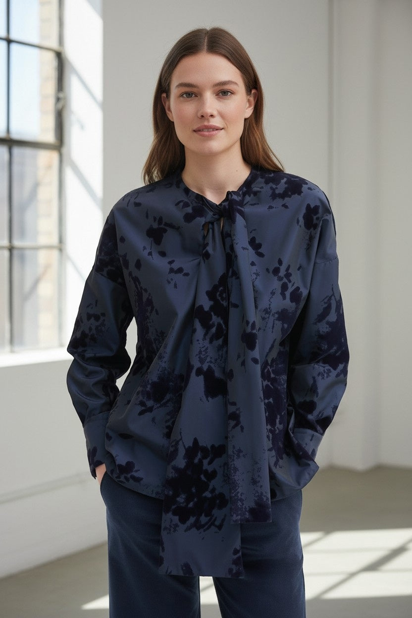 A woman with long brown hair stands indoors by a window, smiling softly with her hands in her pockets. She is wearing the ROSSO35 Taffetta Blouse with Bow Neckline and Velvet by ROSSO 35 SILKY, paired with matching pants.
