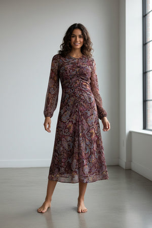 A woman stands facing away, wearing the MIRTO 1956 Burgundy Draped Paisley Print Dress—a long sleeve midi dress featuring an elegant paisley pattern—paired with burgundy tights and black heels. Her hair is styled in a neat bun as she poses against a plain white background.