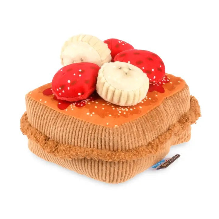Close-up of the PLAY PET LIFESTYLE THICK ‘N FLUFFY FRENCH TOAST DOG TOY on a brown fabric with red and white patterns, pictured with plush strawberry and muffin toys.