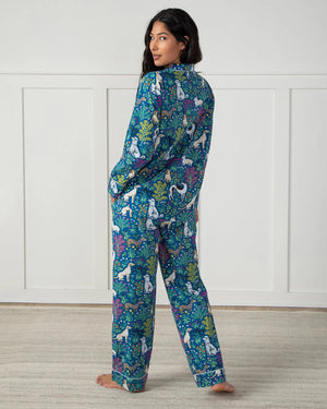 A woman in PRINTFRESH's colorful "Must Love Dogs" organic cotton pajama set stands barefoot on a light rug against a white paneled wall. Her dark hair falls freely as she casually places her hands in her pockets, facing slightly to the side.