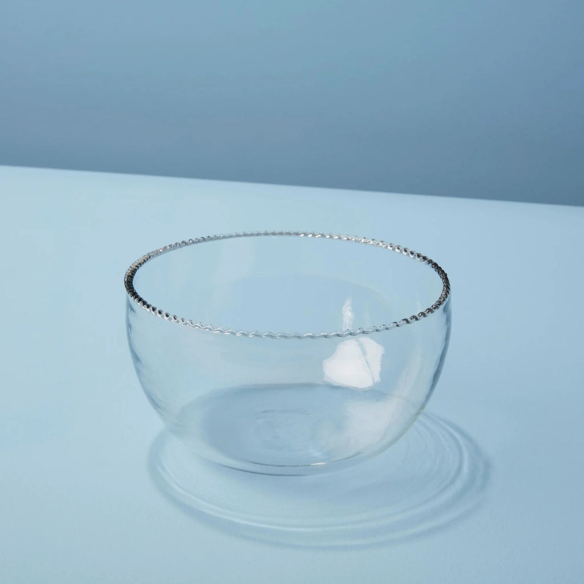 The RUFFLE GLASS BOWL SMALL by BE HOME, INC sits elegantly on a white surface against a pale blue background. The simple, transparent design of this clear glass bowl, featuring a delicate and textured rim, allows the light to cast soft reflections and shadows, creating a clean and minimalist aesthetic.