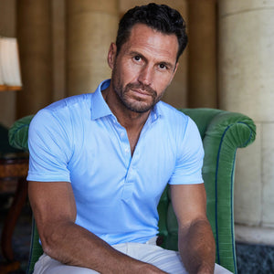 A man with short dark hair and a trimmed beard wears the COLLARS AND CO - SEMI SPREAD COLLAR POLO BLUE HOUNDSTOOTH, paired with light-colored pants. He stands indoors, leaning against a column with a neutral expression.