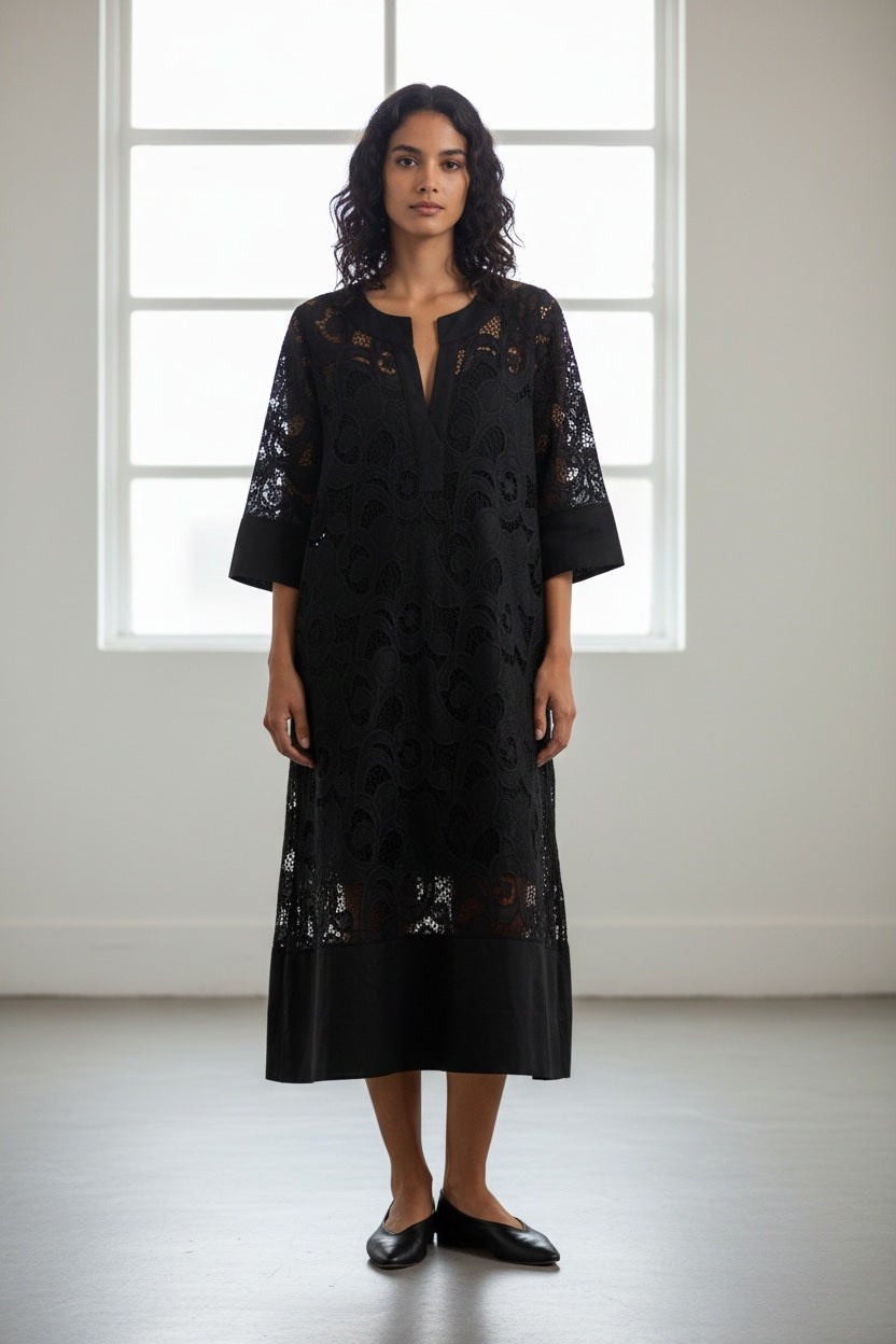 A woman in the MILVA MI Embroidered Cotton Dress in Black by MILVA, featuring three-quarter sleeves and subtle embroidery, stands against a white background. She has short hair, sunglasses, white earrings, and flat shoes.