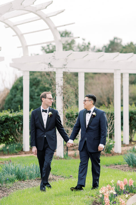 Two men in tuxedos hold hands while walking through a garden with white pergolas and greenery. They look at each other, both smiling gently, surrounded by blooming flowers and natural beauty.